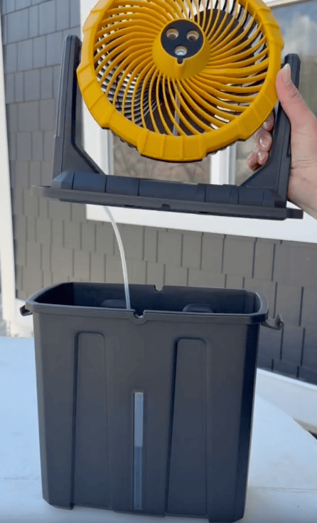 A hand lifts a yellow portable fan above a black rectangular container with a clear water level window, both placed on a white outdoor surface in front of a gray building.