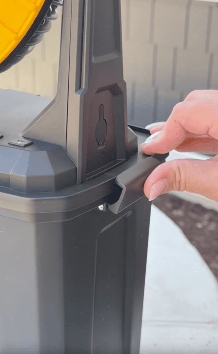 A hand unlatches a gray plastic container with a yellow lid outdoors, near a concrete surface and brown mulch.