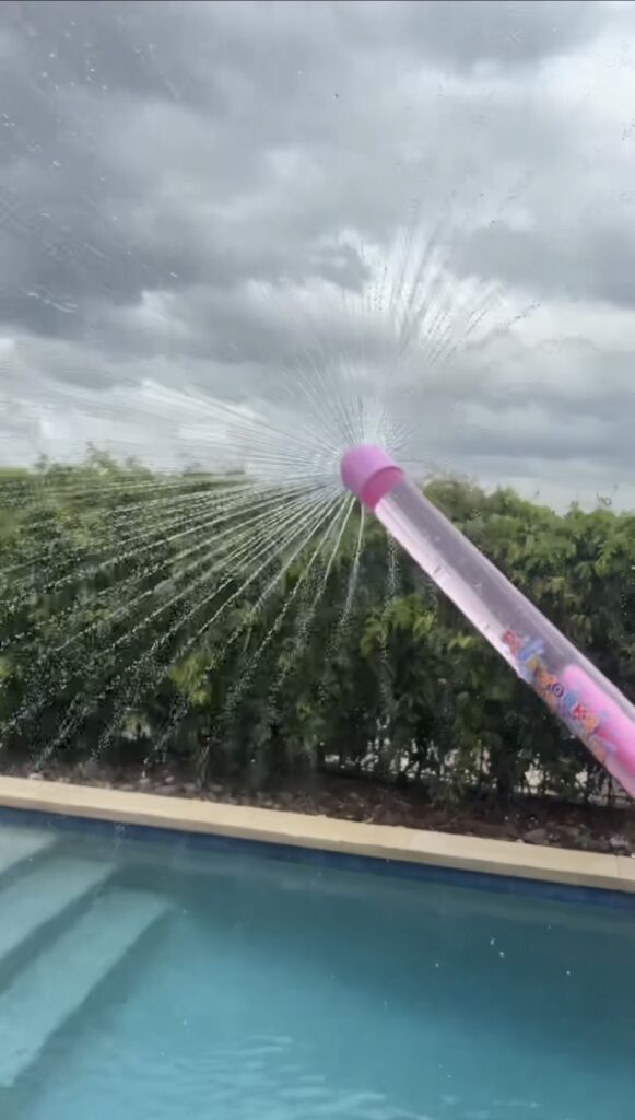 A pink water blaster sprays water in a fan shape near a swimming pool, with green bushes and a cloudy sky in the background.