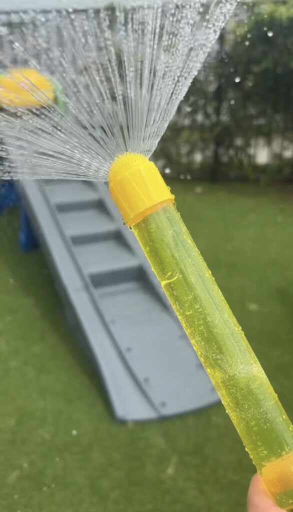 A hand holds a yellow water toy spraying water in a fan shape, with a playground slide and green grass visible in the background.