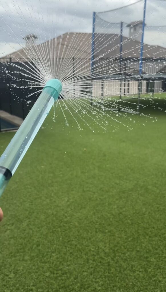 A person holds a blue garden hose nozzle spraying water in a radial pattern over a green lawn, with a trampoline and house visible in the background.