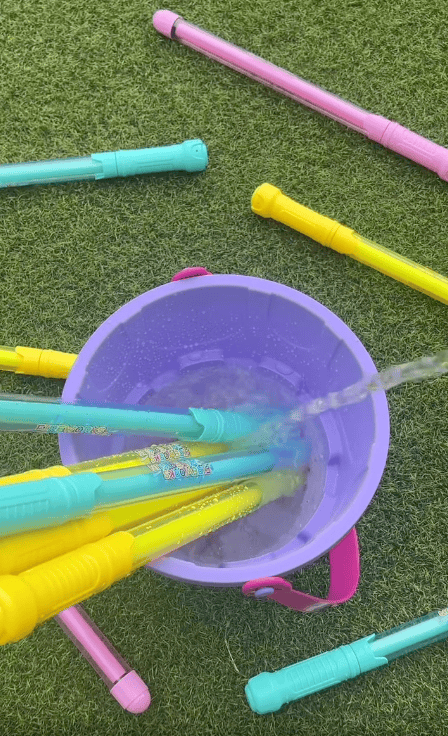 Colorful bubble wands are placed inside a purple bucket while water flows into it, with more wands scattered on green artificial grass around the bucket.
