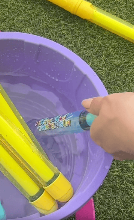 A hand holds a blue and yellow water gun labeled "Fireworks Water Gun," dipping it into a purple bucket filled with water and other water guns. The scene is set on green grass.