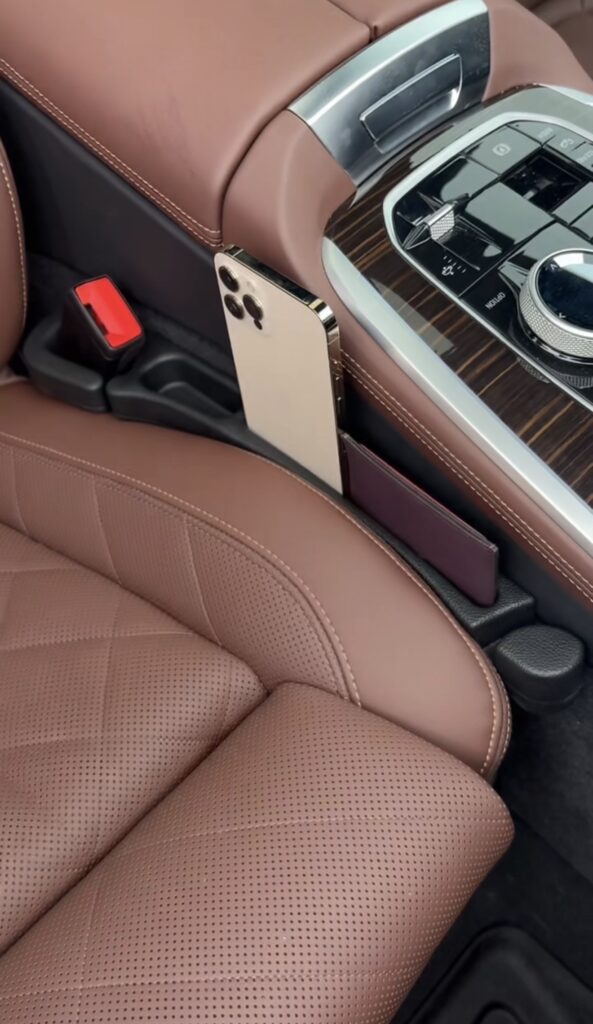A gold smartphone and a slim wallet are placed in a gap filler organizer between a brown leather car seat and the center console inside a vehicle.