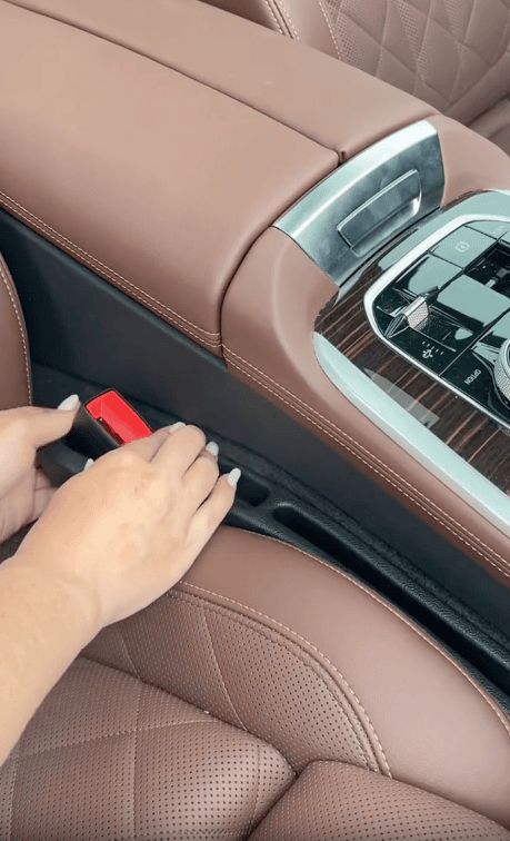 A person inserts a black gap filler strip between the brown leather car seat and the center console to prevent items from falling into the gap.