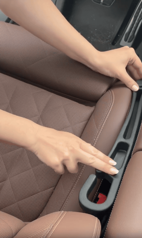 A person places a black car seat gap filler between a brown car seat and the center console, using their fingers to adjust it into position.