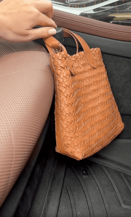 A hand secures a tan woven leather handbag to a car seat using a black strap, with the bag resting on the car's floor mat beside the brown perforated seat.