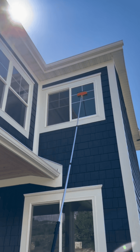 A long-handled brush is being used to clean the exterior window of a two-story house with dark blue siding under a sunny, clear sky.