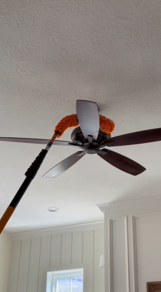 A person uses an orange microfiber duster on an extendable pole to clean the blades of a ceiling fan in a room with white walls and a textured ceiling.