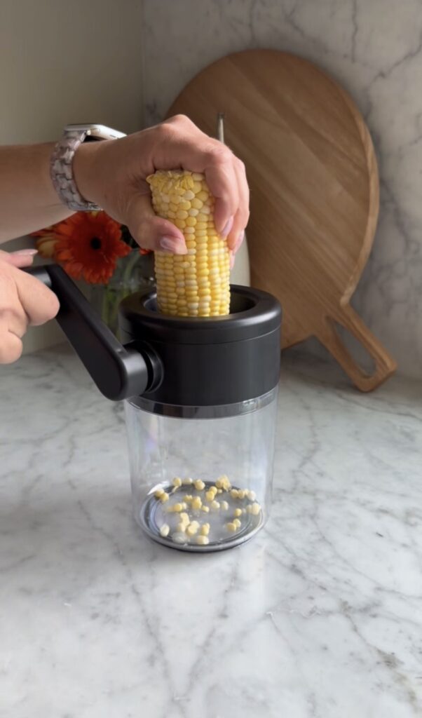 A person uses a hand-cranked corn peeler to remove fresh corn kernels from a cob, collecting them in a clear container on a marble countertop, with a wooden cutting board and flowers in the background.