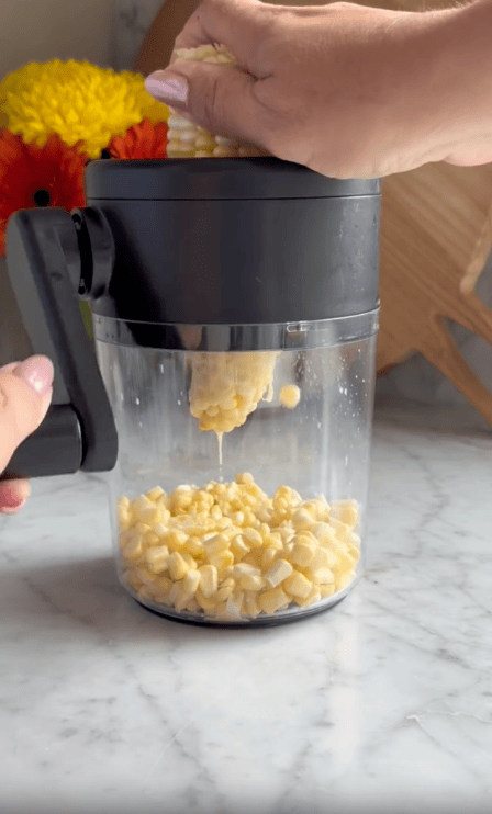 A person uses a corn peeler to remove fresh corn kernels from the cob, collecting them in a clear container with a black lid, set on a marble countertop with flowers and a wooden cutting board in the background.
