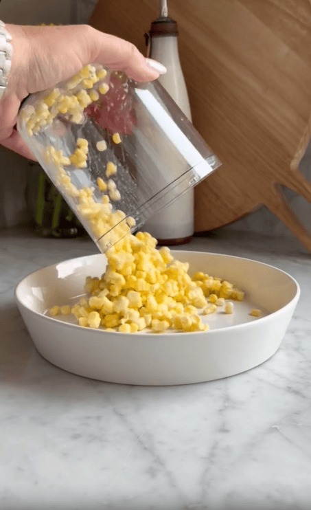 A hand pours a container of fresh corn kernels into a shallow white dish on a marble countertop, with a wooden cutting board and bottle in the background.