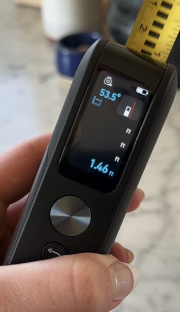 A hand holds a digital measuring tape showing a screen with a 53.5° angle, a low battery icon, and measurements of 1.46 feet and 5 feet. The device is dark-colored with a round silver button.