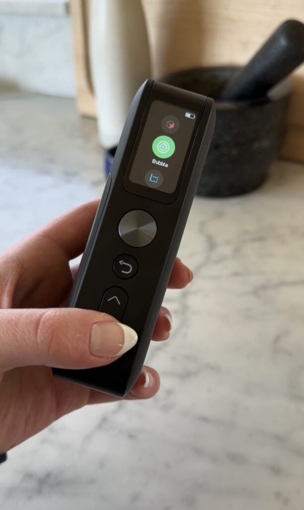 A hand holding a black electronic device with a screen displaying the word "Saúde" and icons. The device has buttons below the screen and is held over a marble countertop.