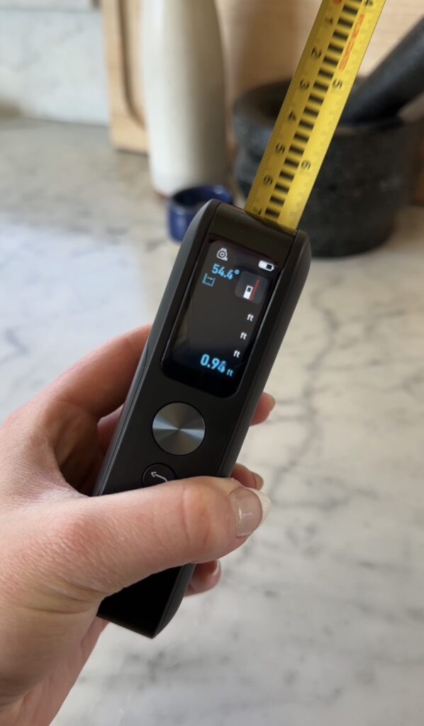 A hand holds a digital tape measure displaying 5.41 ft and 0.95 ft on its screen. The yellow tape is extended, and the background shows a marble surface and blurred objects.