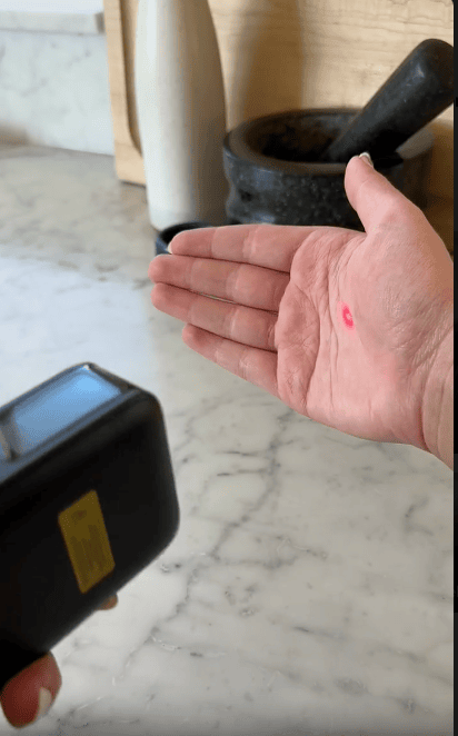 A hand with a red laser dot on the palm is held open over a marble countertop, while a person points a handheld scanner device at the palm. A mortar and pestle are visible in the background.