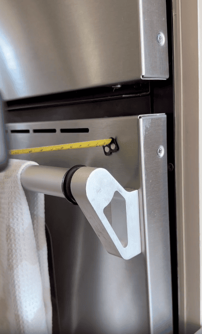 A close-up of a metal towel holder attached to a stainless steel appliance door, with a yellow measuring tape stretched across the opening.