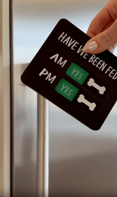 A hand holds a sign with switches labeled "Have we been fed? AM Yes, PM Yes" and dog bone icons, used to track pet feeding times.