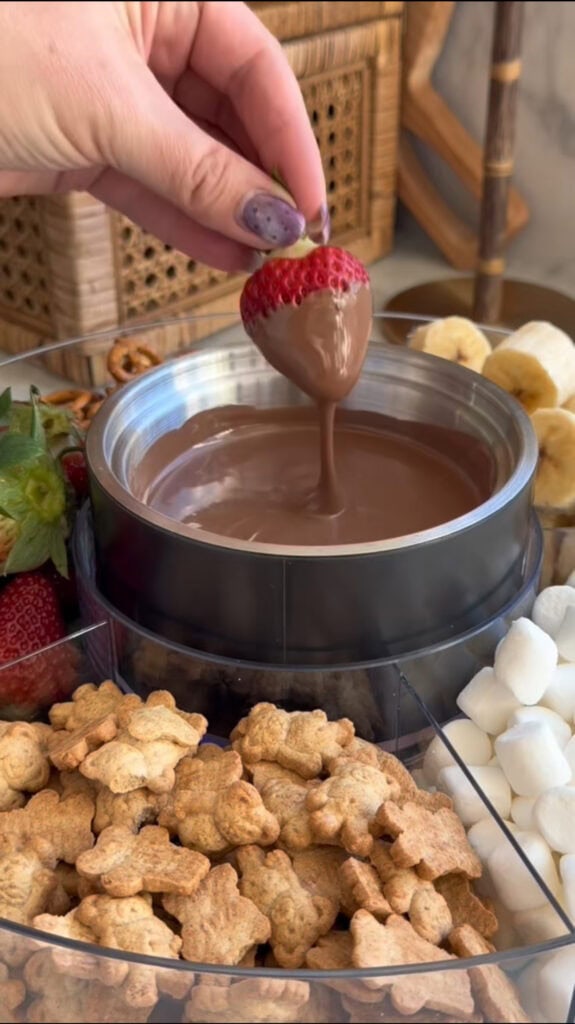 A hand dips a strawberry into a bowl of melted chocolate, surrounded by bear-shaped cookies, marshmallows, banana slices, and more strawberries on a platter.
