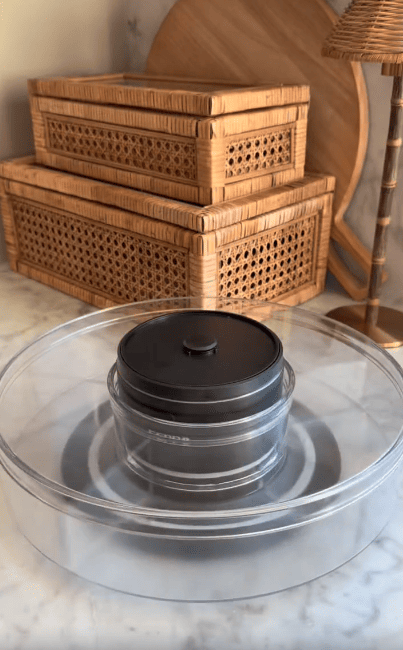 A clear plastic lazy Susan holds a black container with a lid, placed on a marble surface. Behind it are two stacked wicker boxes and a decorative lamp.