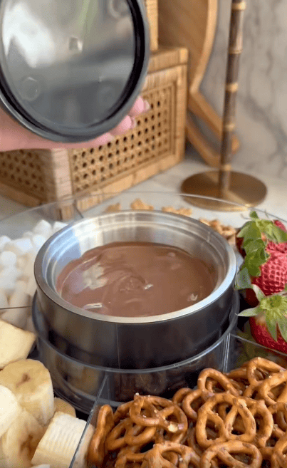 A hand lifts the lid off a container of melted chocolate surrounded by banana slices, strawberries, and pretzels on a tray, ready for dipping.