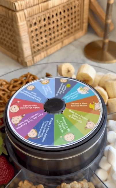 A close-up of a spinning wheel game with colorful sections, surrounded by snacks like pretzels, bananas, strawberries, and marshmallows on a tray.