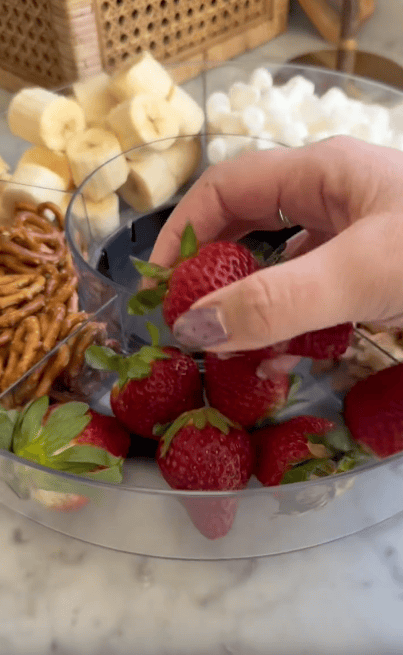 A hand picks up a strawberry from a divided tray containing strawberries, pretzels, banana slices, and marshmallows on a marble countertop.