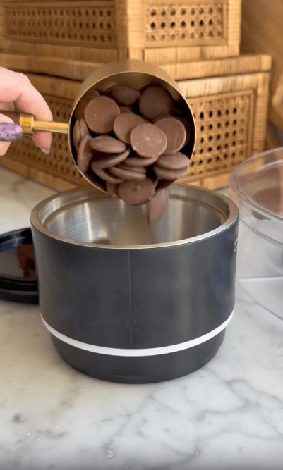A hand pours chocolate wafers from a gold measuring cup into a black and silver container on a marble countertop, with wooden boxes in the background.