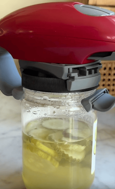 A red automatic jar opener is gripping the lid of a glass jar filled with pickle slices and brine on a light-colored countertop.