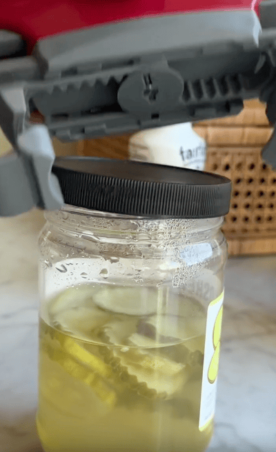 A jar opener grips the lid of a pickle jar, preparing to twist it open. The jar is on a countertop with a wicker basket in the background.