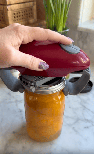 A hand uses a red automatic jar opener to open a glass jar filled with orange contents on a marble countertop.