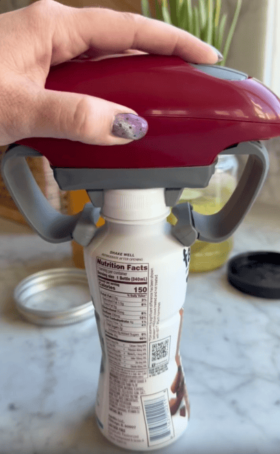A hand uses a red and gray jar opener to grip a plastic bottle with visible nutrition facts. The scene is set on a kitchen counter with blurred items in the background.
