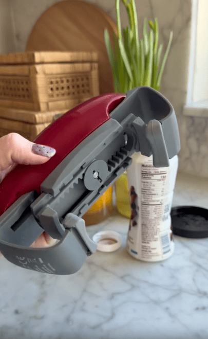 A hand holds a gray and red jar opener above a marble countertop with a can, a black lid, wooden containers, and green onions in the background.