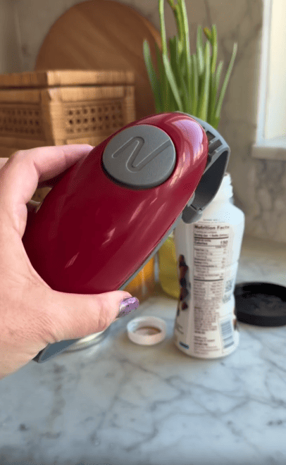 A hand holds a red and gray electric can opener over a white can on a marble countertop, with kitchen items and green onions in the background.