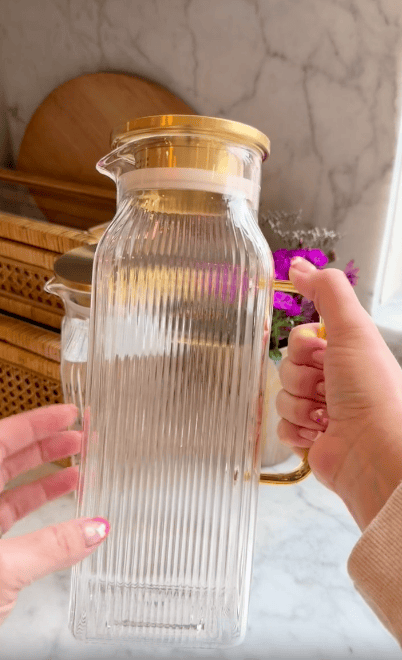A hand holds a tall, clear, ribbed glass pitcher with a gold lid and handle. Another hand is reaching toward it. The background shows kitchen items and a vase with purple flowers.