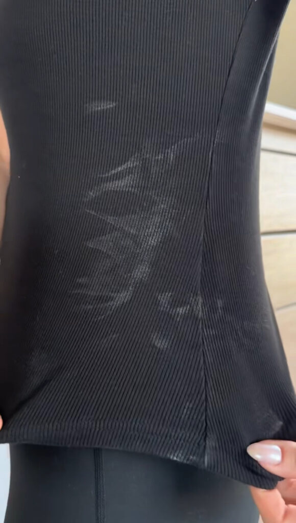 A close-up of a person holding the bottom of a black ribbed shirt, showing visible white deodorant stains on the fabric.