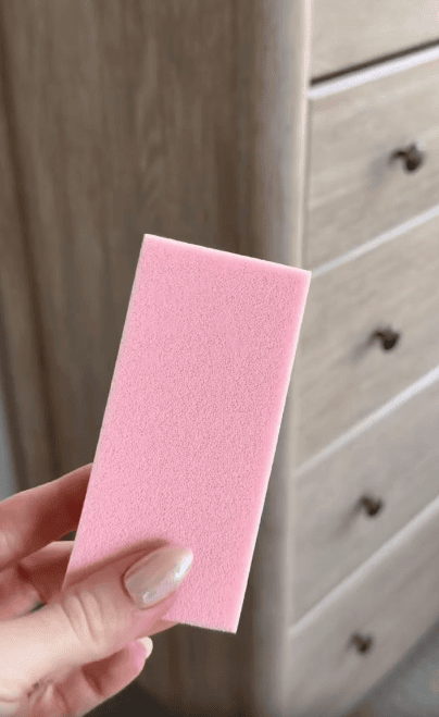 A hand with light-colored nails holds a pink rectangular sponge in front of a wooden dresser with several drawers.