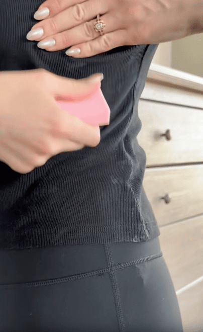 A person uses a pink pumice stone to remove lint from a black ribbed shirt. Their hand with manicured nails and a ring is resting on their side. A wooden dresser is in the background.