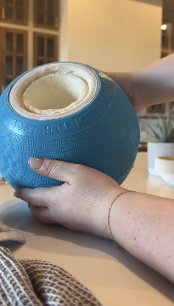 A person holds a blue softshell ice cream maker with freshly made ice cream inside, resting on a kitchen counter near a towel and a small potted plant.