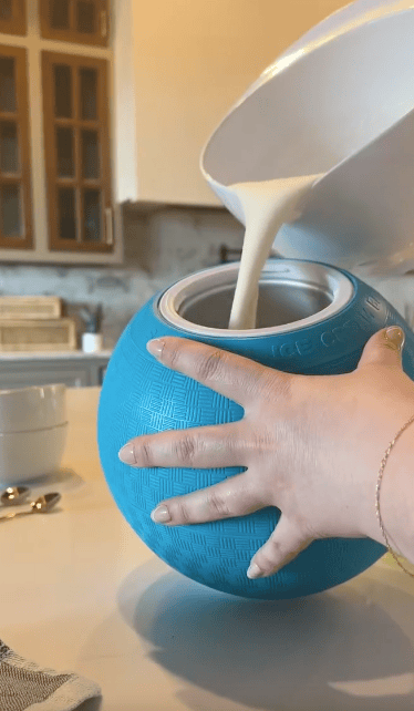 A hand holds a blue ice cream maker ball while a thick white mixture is poured into it from a bowl in a kitchen setting.