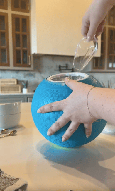 A person pours water into a blue ball-shaped ice cream maker on a kitchen counter, holding it steady with one hand.