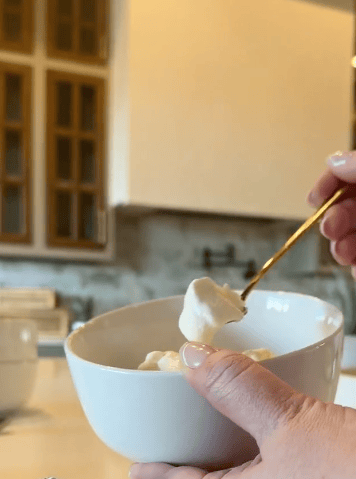 A hand holds a white bowl with marshmallows, while another hand uses a gold spoon to scoop one out. A kitchen with cabinets is blurred in the background.