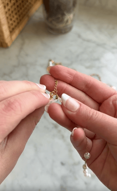 Close-up of hands with French manicured nails fastening a clasp on a delicate gold bracelet with pearl-like beads on a marble surface.
