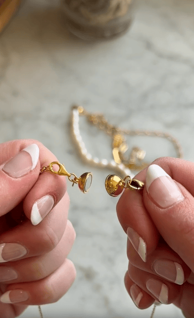 Two hands with French manicured nails hold the gold clasp and loop of a necklace, preparing to fasten it. A blurred gold chain with pearls is visible on a marble surface in the background.