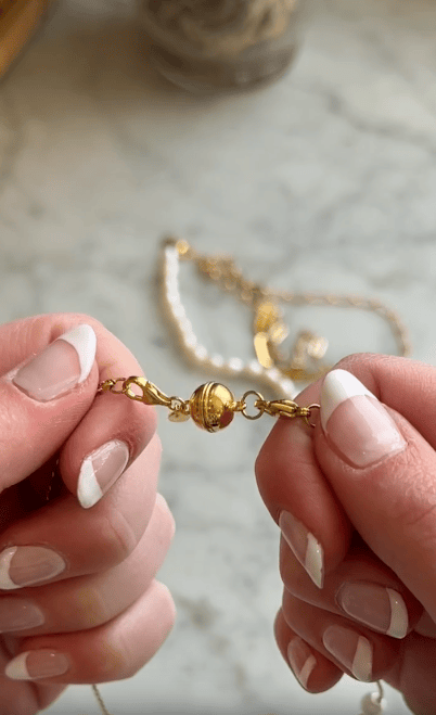 Two hands with French manicured nails hold a gold necklace with a ball clasp, against a blurred marble surface with another necklace in the background.