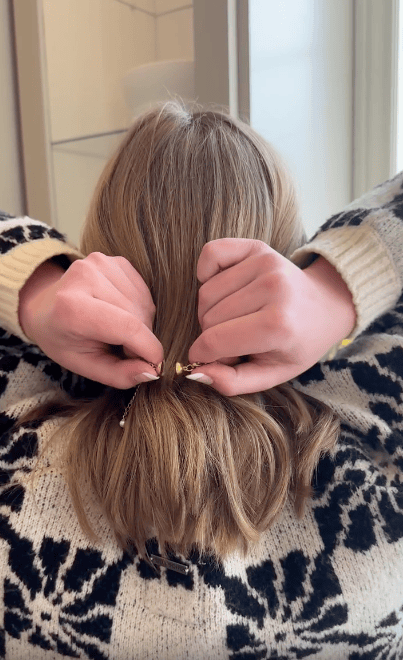 A person with light brown hair and a patterned sweater uses both hands to fasten a necklace behind their neck, viewed from the back.