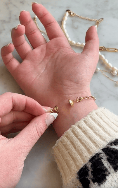 A hand is fastening a gold bracelet with a small heart charm around another wrist. A pearl and gold chain bracelet lies on a marble surface in the background.