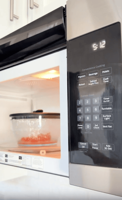 A microwave with the door slightly open shows food in a covered dish inside. The display reads 5:12, and the keypad includes options like popcorn, beverage, and timer settings.