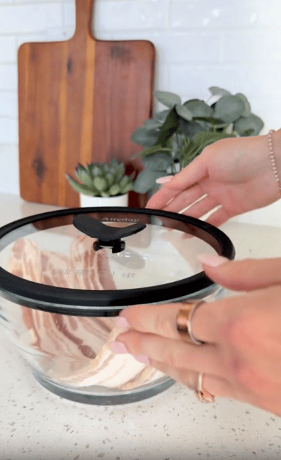 A person places a glass lid on a bowl containing raw bacon strips, with a wooden cutting board and plants in the background on a white kitchen counter.