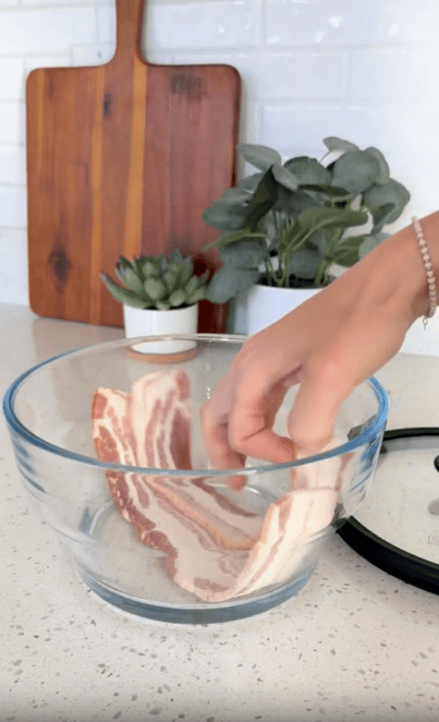 A hand places raw bacon strips into a clear glass bowl on a kitchen counter, with a cutting board, plants, and kitchen items in the background.